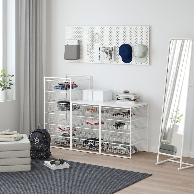 a white shelf with baskets and clothes on it in front of a large mirror, next to a gray rug