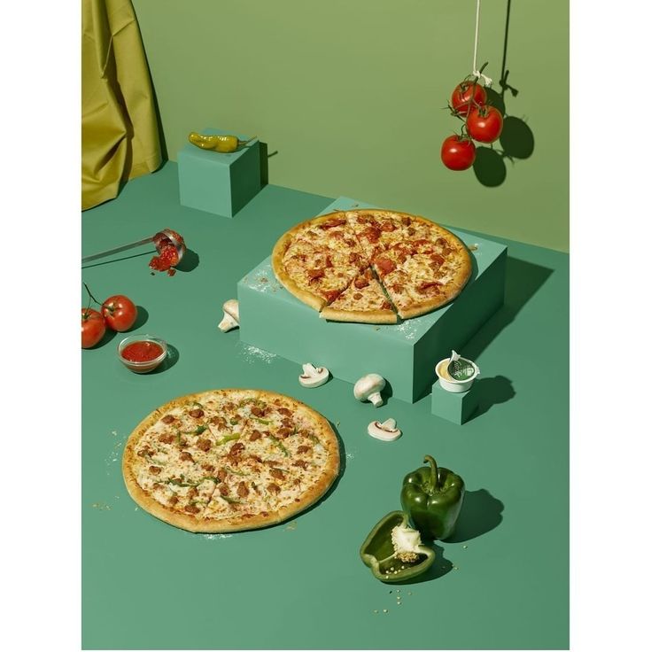 two pizzas sitting on top of a green table next to peppers and tomatoes, with one slice missing