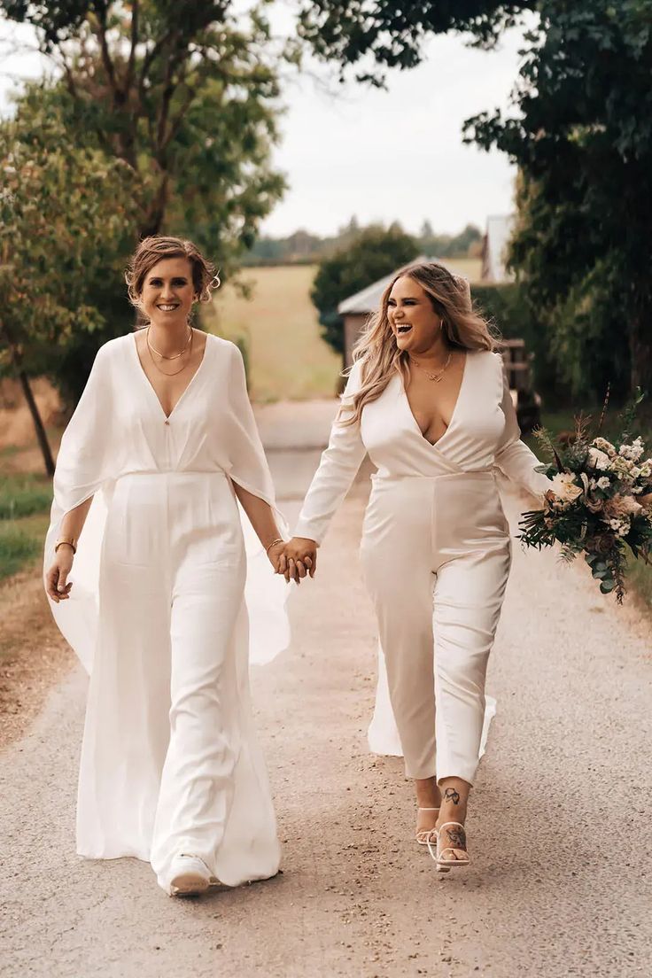 two women in white outfits walking down a dirt road holding hands and smiling at the camera