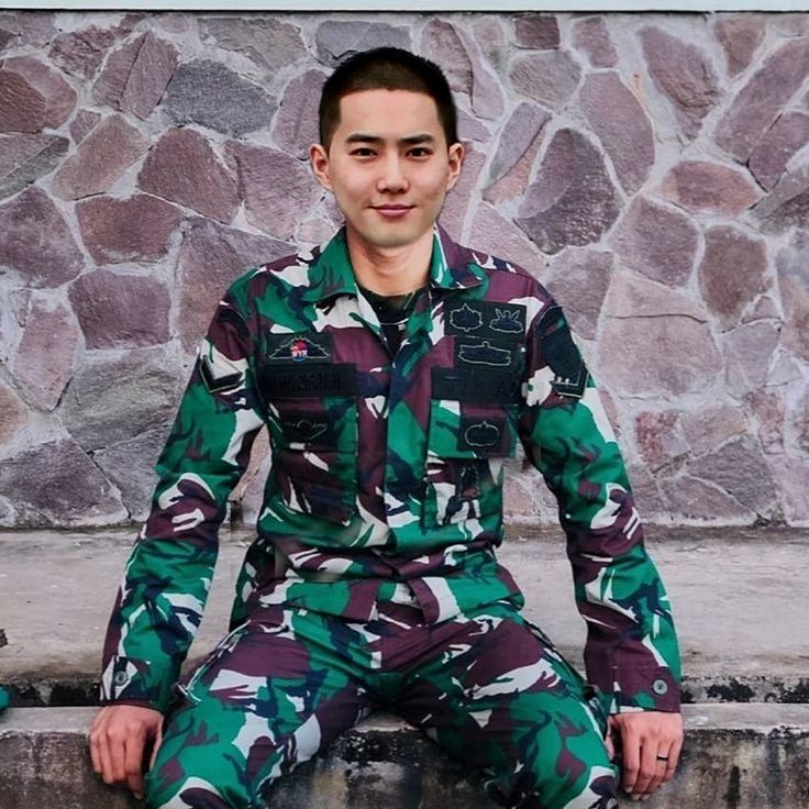 a young man sitting on the ground wearing camouflage clothing with his hands in his pockets