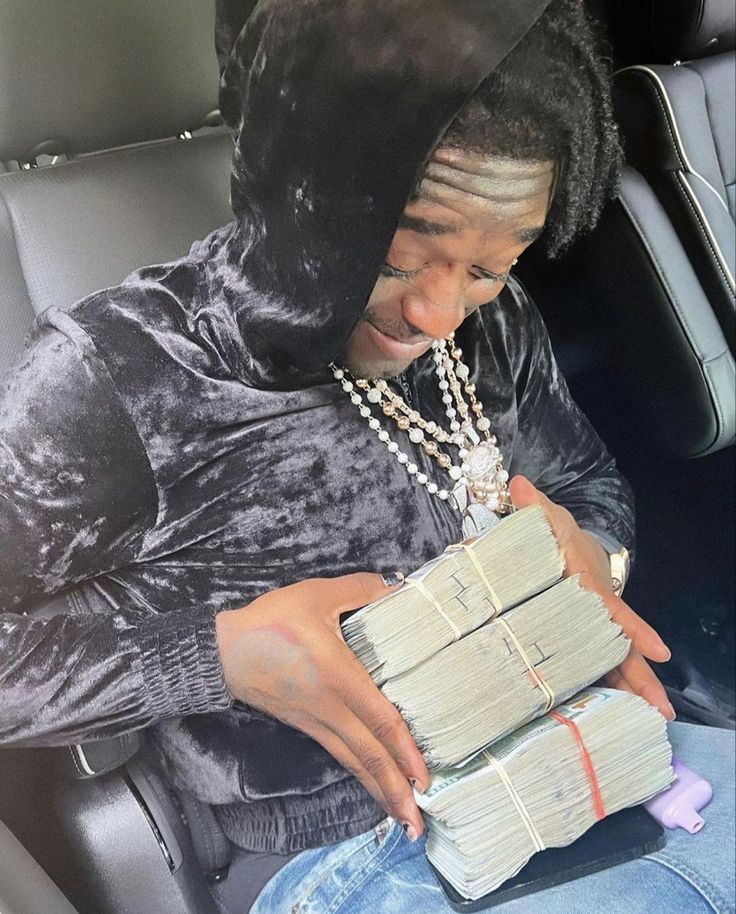 a man sitting in the back seat of a car holding stacks of money and wearing a hoodie