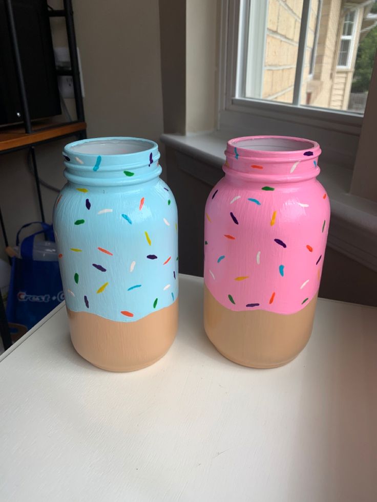 two painted jars with sprinkles on them sitting in front of a window