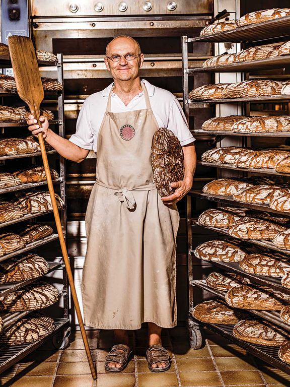 an older man holding a wooden spatula in front of a rack of baked goods