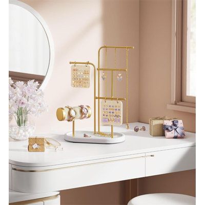 a white desk topped with a mirror and gold jewelry holder next to a vase filled with flowers