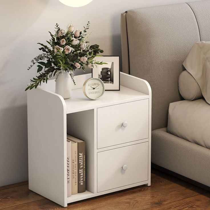 a white nightstand with flowers and books on it next to a couch in a living room