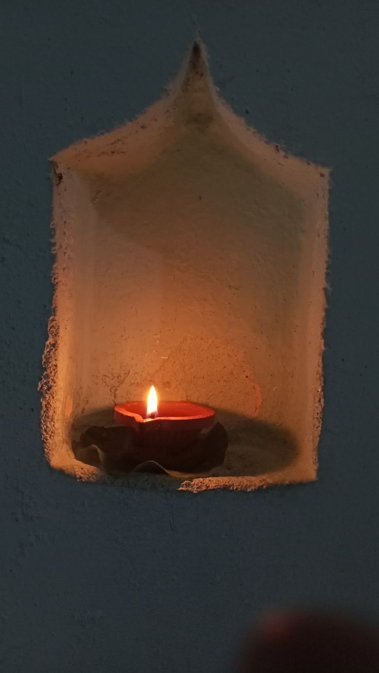 a lit candle in a white wall with a small hole between it and someone's hand