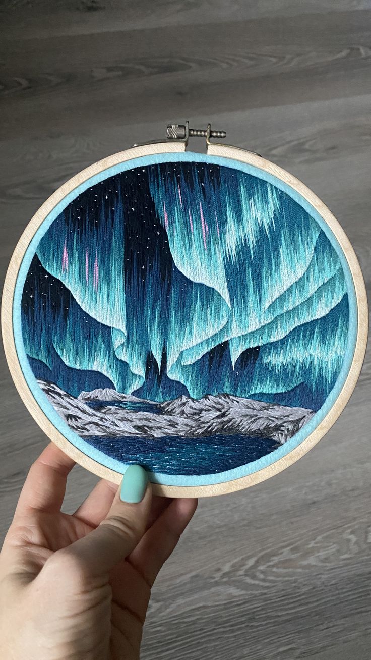 a hand holding up an embroidery project with aurora bores in the sky on it