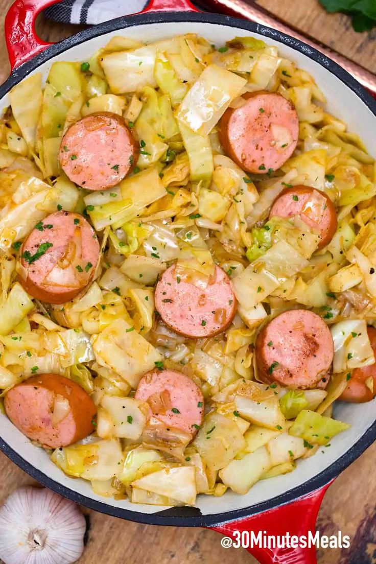 sausages and cabbage in a skillet on a wooden table with garlic, pepperoni, and onions