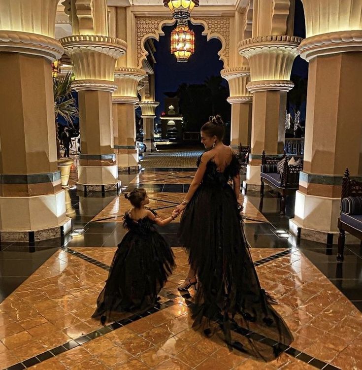 two women in black dresses are walking through an ornate hall with columns and chandeliers