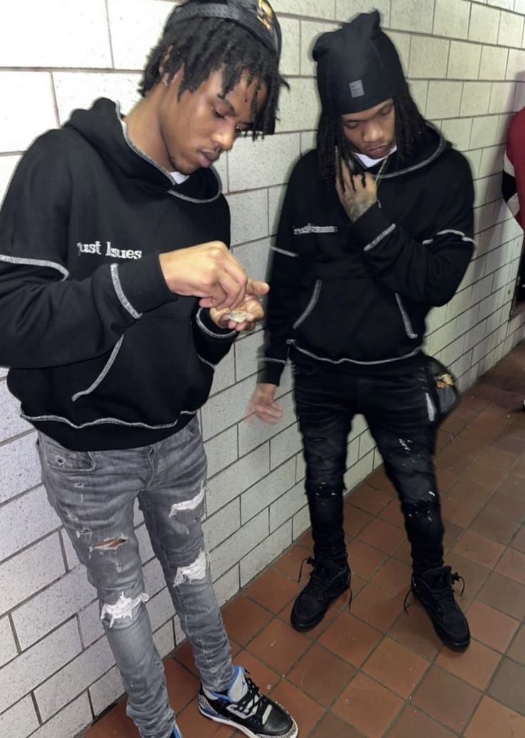 two young men standing next to each other in front of a brick wall looking at their cell phones