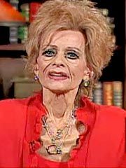 an older woman in a red jacket is sitting down and smiling at the camera, with books behind her