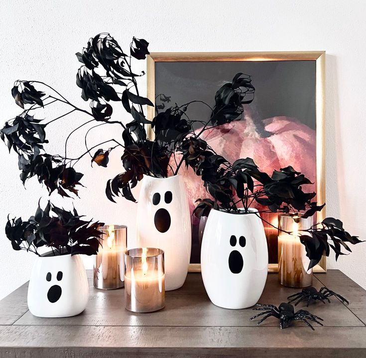 some white vases filled with plants and candles on top of a wooden table in front of a painting