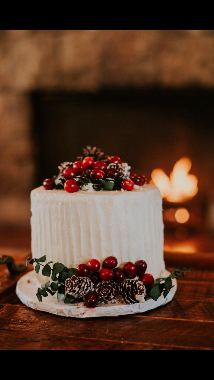 a white cake with berries and pine cones on top sits on a table in front of a fireplace