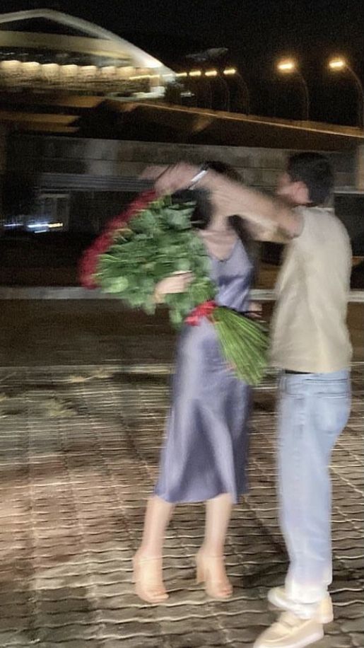 a man and woman standing next to each other on a brick walkway at night, holding flowers