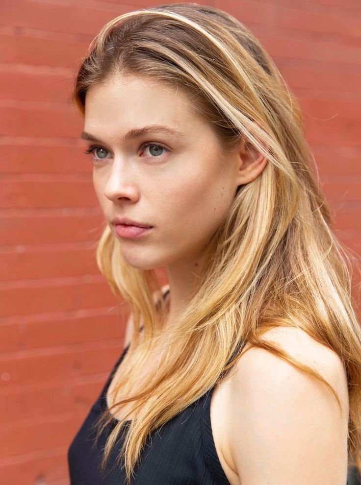 a woman with long blonde hair standing in front of a brick wall and looking at the camera