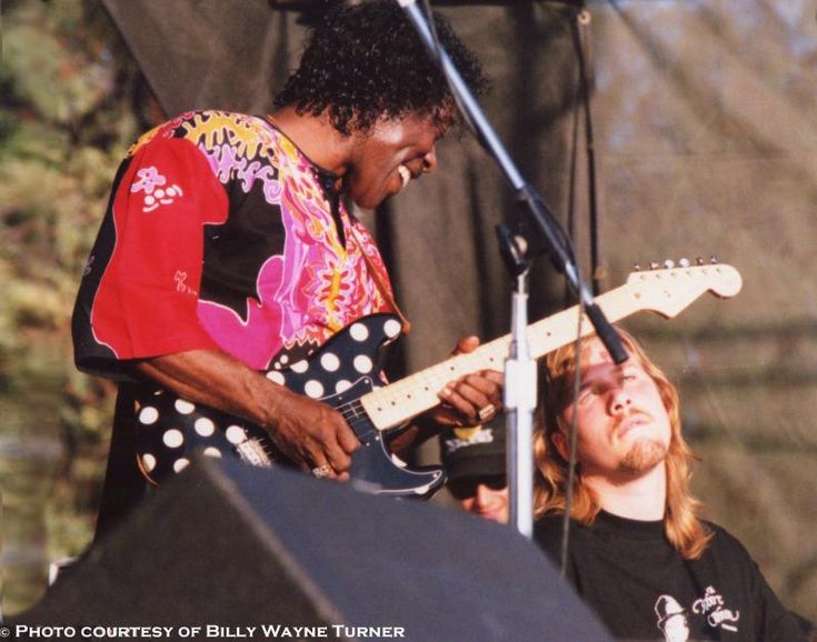 two men playing guitars on stage at an outdoor concert, one man is wearing a colorful shirt and the other wears a black t - shirt with white polka dots