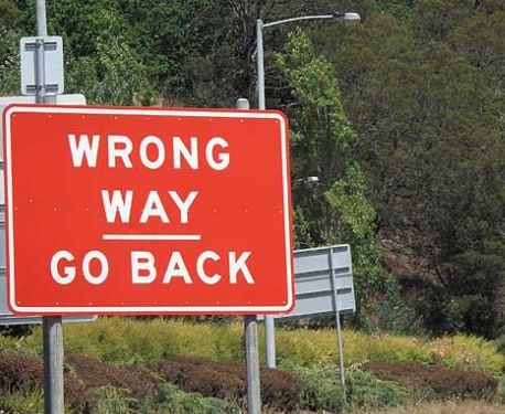 a red sign that says wrong way go back on the side of a road with trees in the background