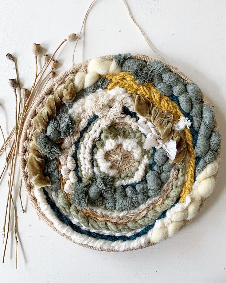 a woven wall hanging made with yarn and cottons, on a white wall next to dried flowers