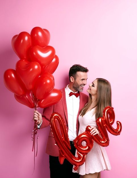 a man and woman standing next to each other holding red balloons in the shape of love