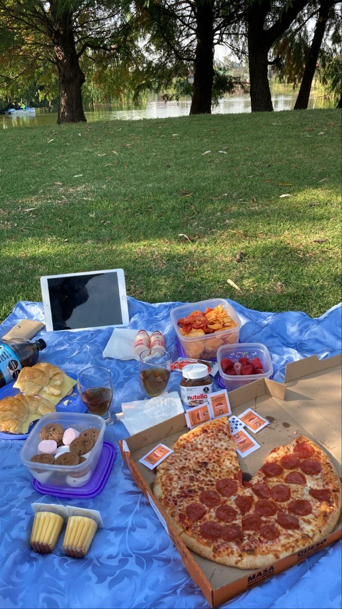 a pizza and other food items on a blue table cloth in the middle of a park