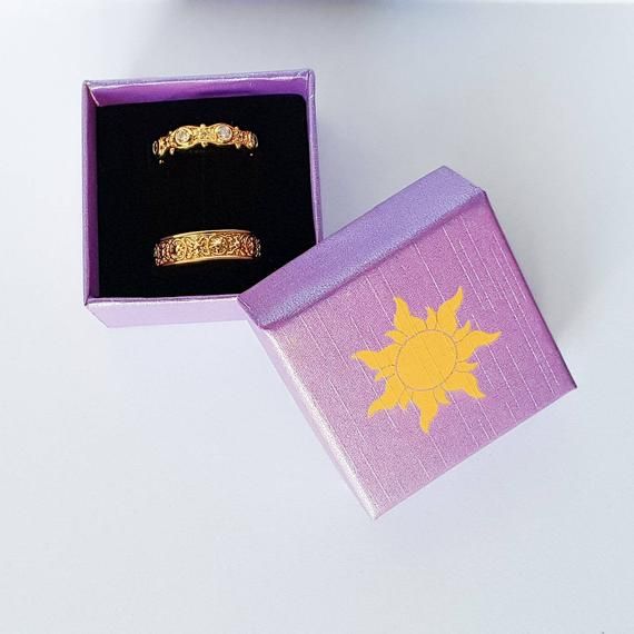 two gold rings in a purple box on top of a white table next to an orange and yellow flower