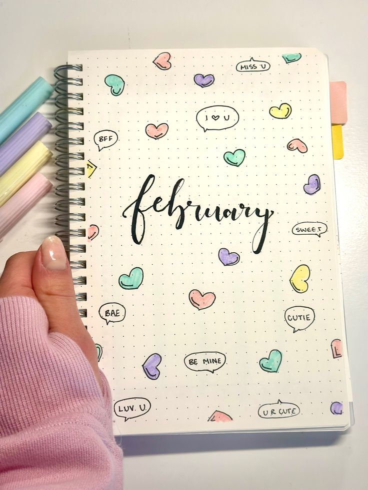 a person holding a notebook with the words february written on it next to colored pencils