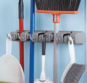 three brooms and two mop hanging from hooks on a blue wall with other cleaning supplies