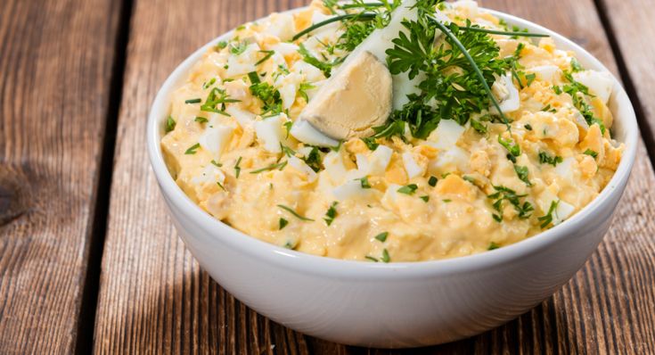 a white bowl filled with eggs and parsley on top of a wooden table next to bread