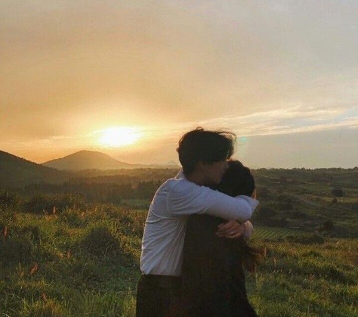 two people embracing each other in a field with the sun setting behind them and hills in the distance