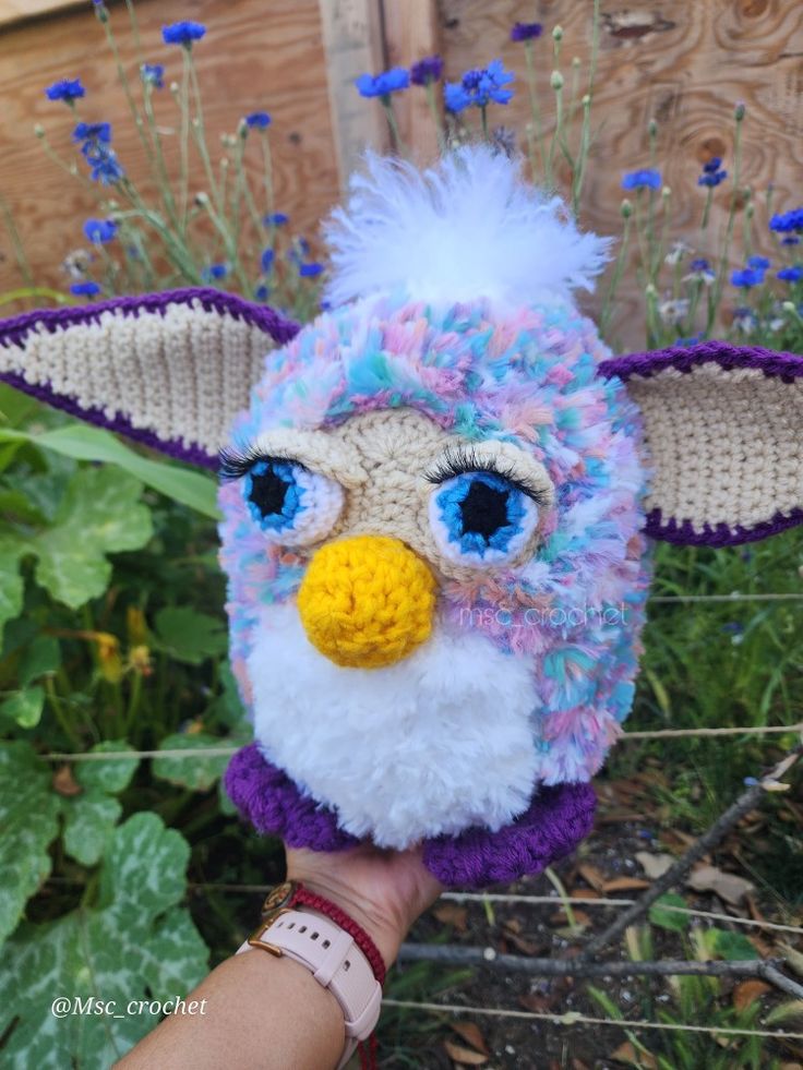 a hand holding a small stuffed animal with blue eyes and purple fur on it's head