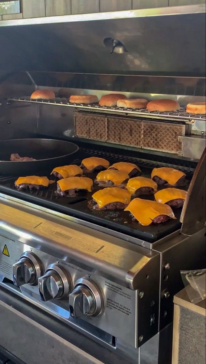 hamburgers being cooked on an outdoor grill with buns in the oven behind them