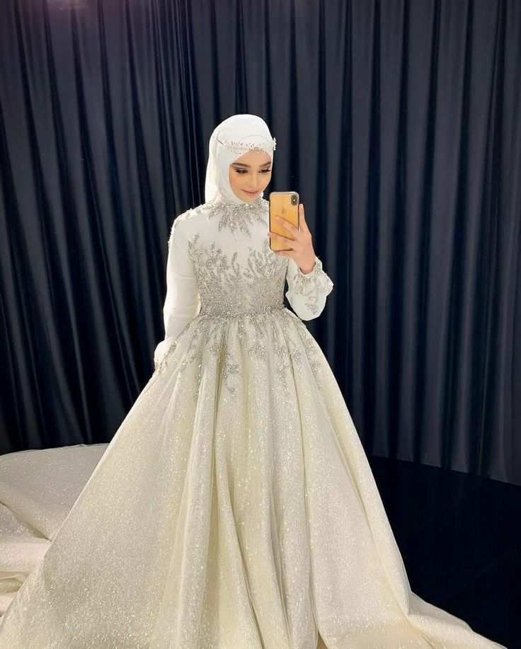 a woman in a white dress taking a selfie with her cell phone while wearing a hijab