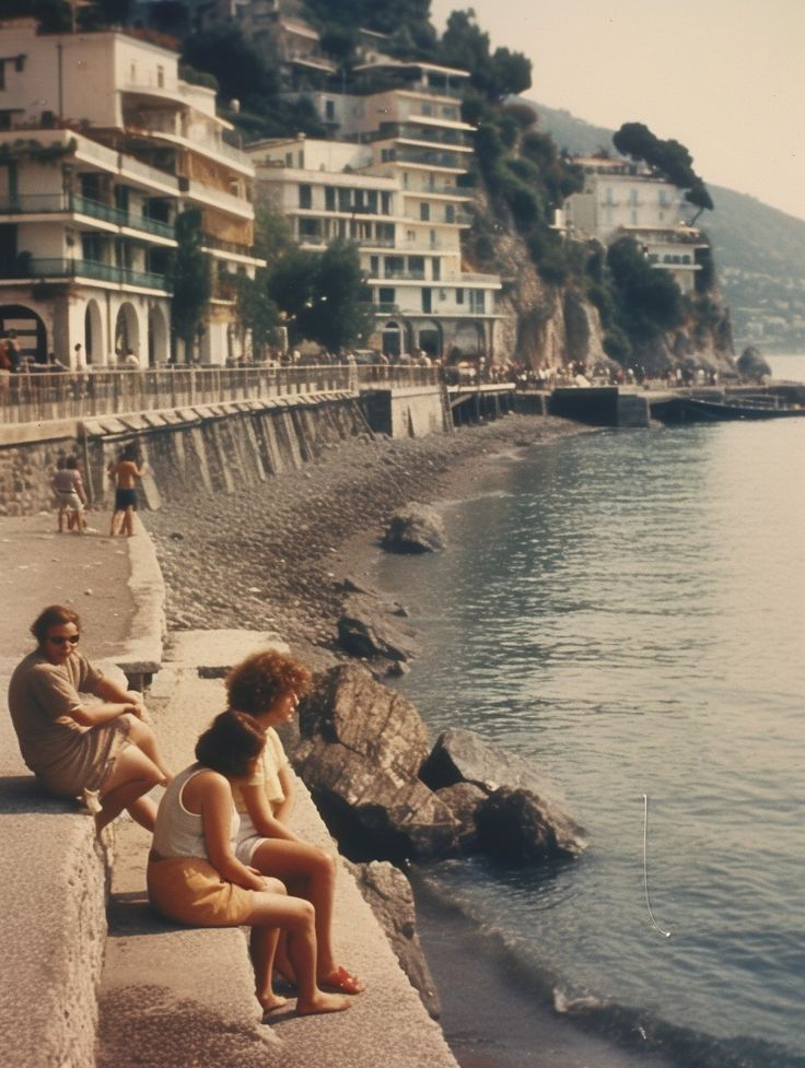 two people sitting on the edge of a body of water next to buildings and cliffs