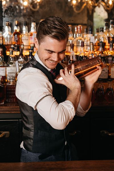 a man in a vest is holding a shaker behind his back at a bar