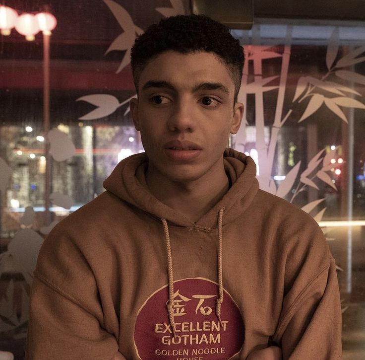 a young man in a brown hoodie is looking at the camera and has his arms crossed