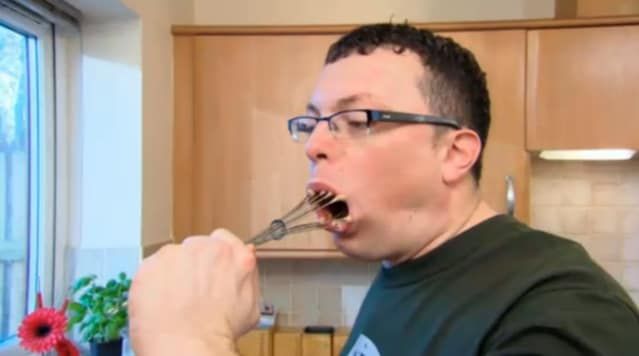 a man with glasses is brushing his teeth in the kitchen while holding a toothbrush up to his mouth