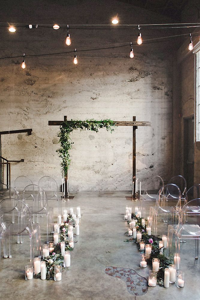 an indoor wedding setup with clear chairs, candles and greenery on the aisle for guests to sit down