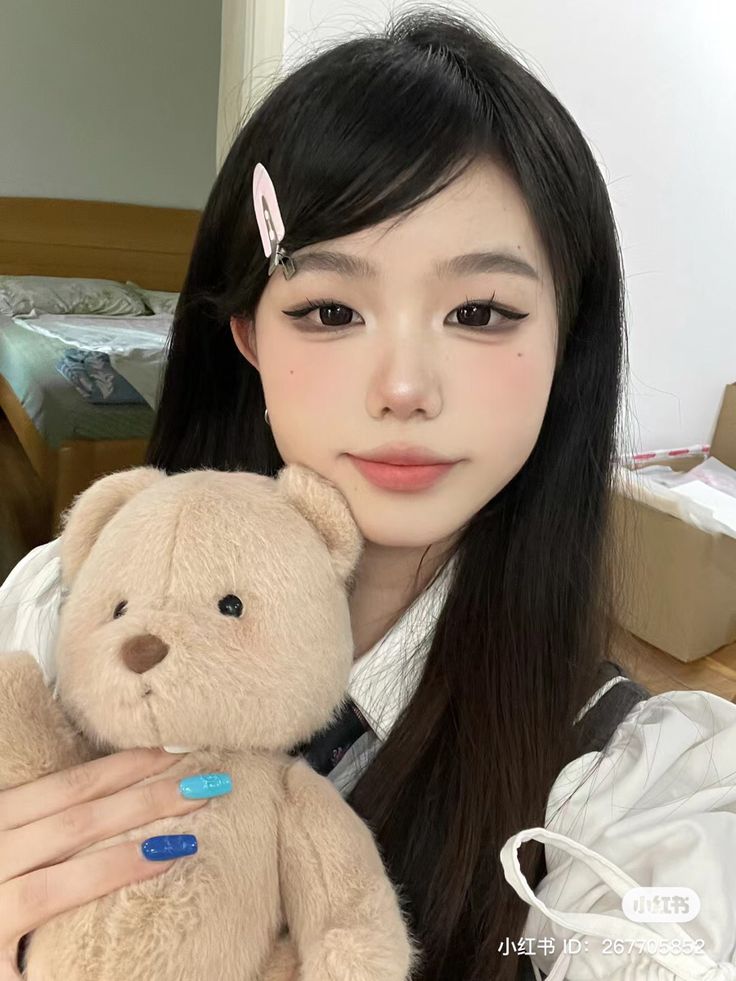 a girl with long black hair holding a brown teddy bear in her right hand and looking at the camera