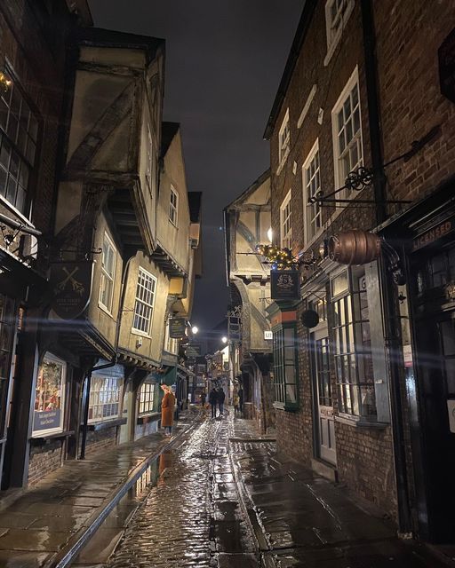 people are walking down an alleyway in the rain with umbrellas and buildings on either side