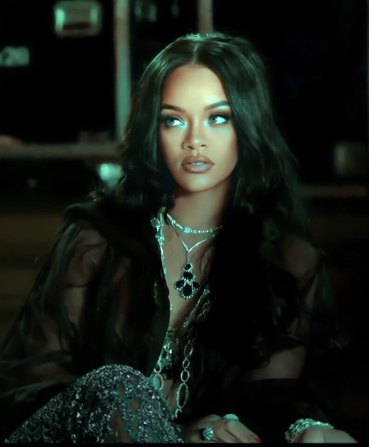 a woman with long black hair sitting down in front of a dark background and wearing jewelry