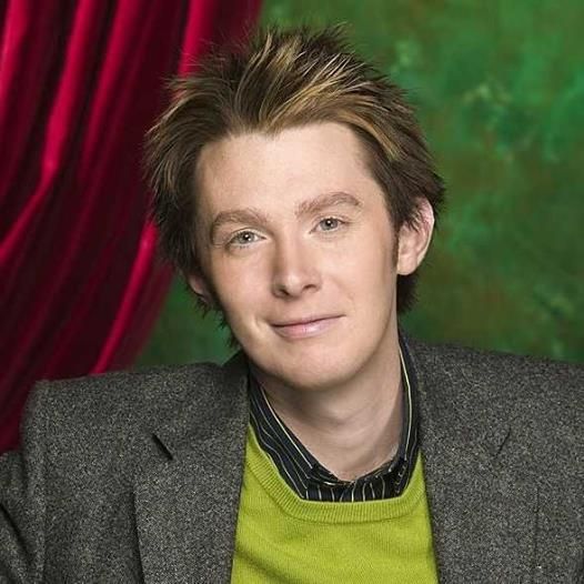 a young man in a gray jacket and green shirt posing for a photo with red curtains behind him