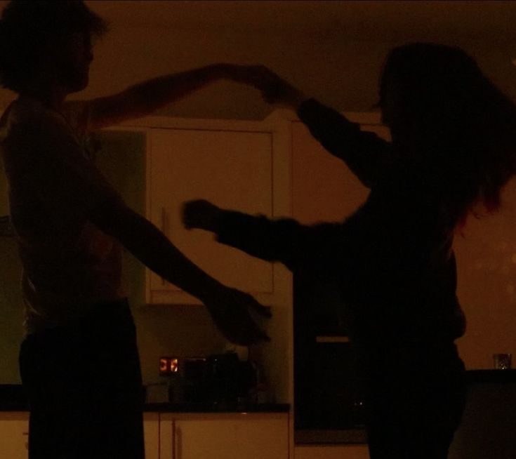 two people are dancing in the kitchen at night, one is holding his arm out