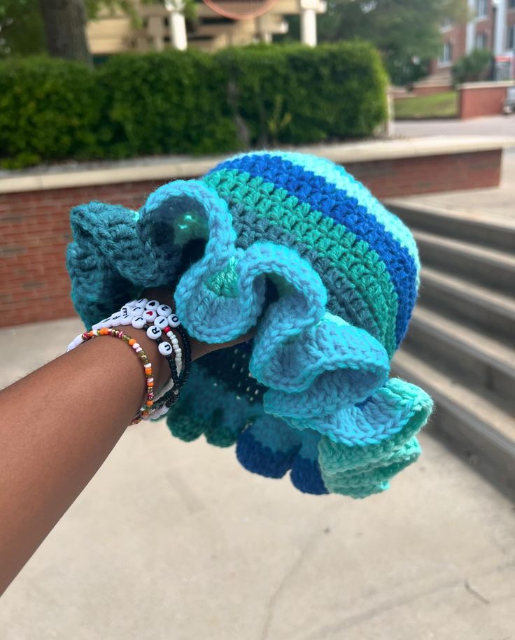 a hand holding up a crocheted blue and green hat on top of concrete steps