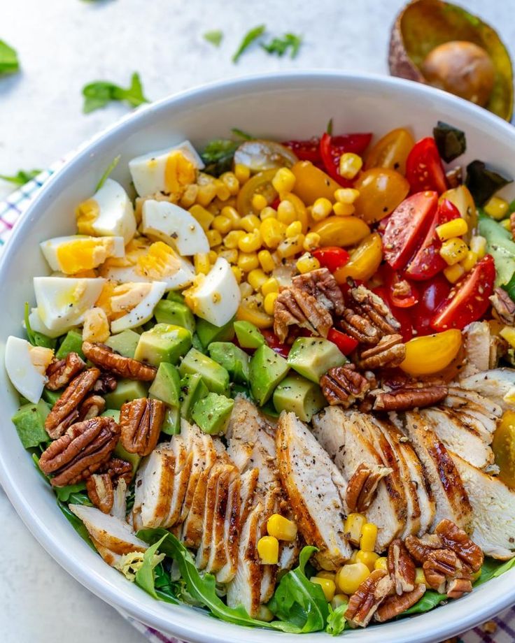 a white bowl filled with chicken, avocado, tomatoes and corn on top of a checkered table cloth