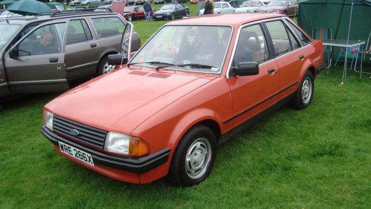 an orange car parked on top of a lush green field next to other cars in the background