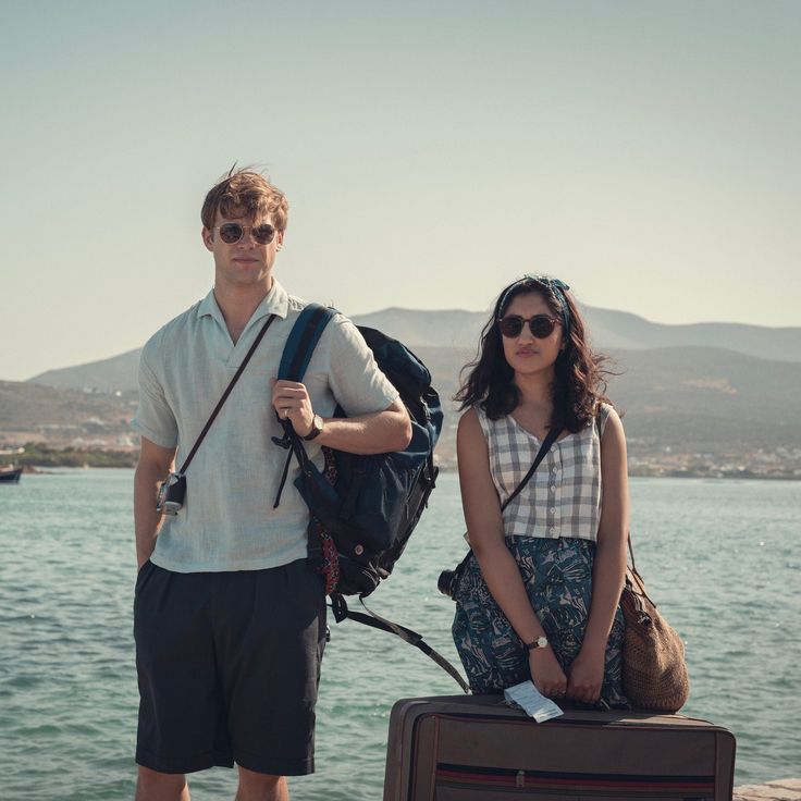 a man and woman standing next to each other by the water with luggage in front of them