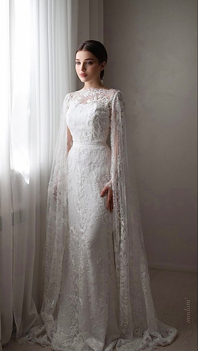 a woman standing in front of a window wearing a white wedding dress with long sleeves