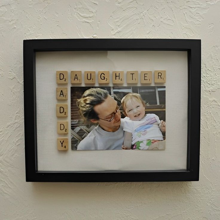 a woman holding a baby in front of a scrabble frame with the word daddy spelled on it