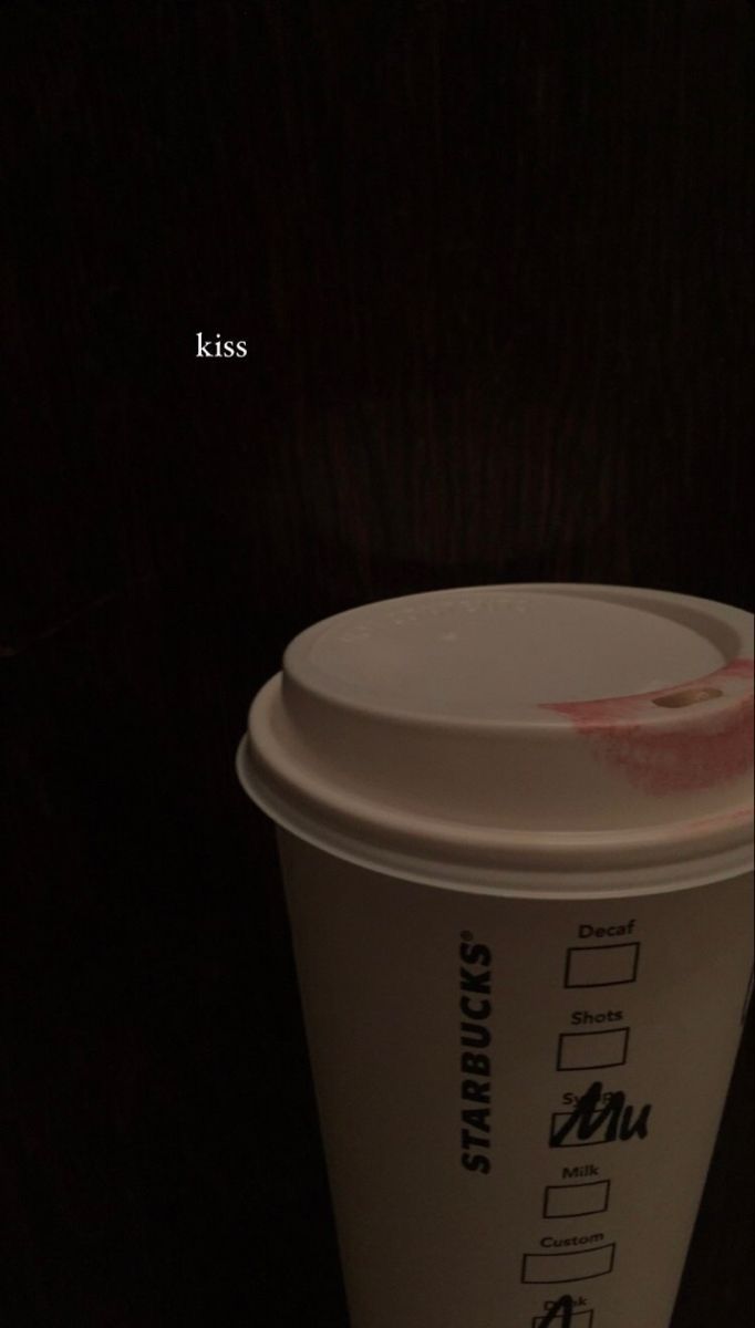 a white cup sitting on top of a wooden table next to a black wall with the words kiss written on it