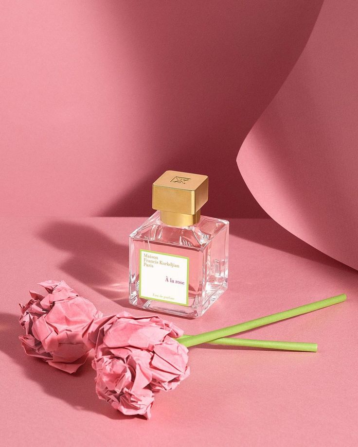 a pink flower next to a bottle of perfume on a pink surface with two green straws